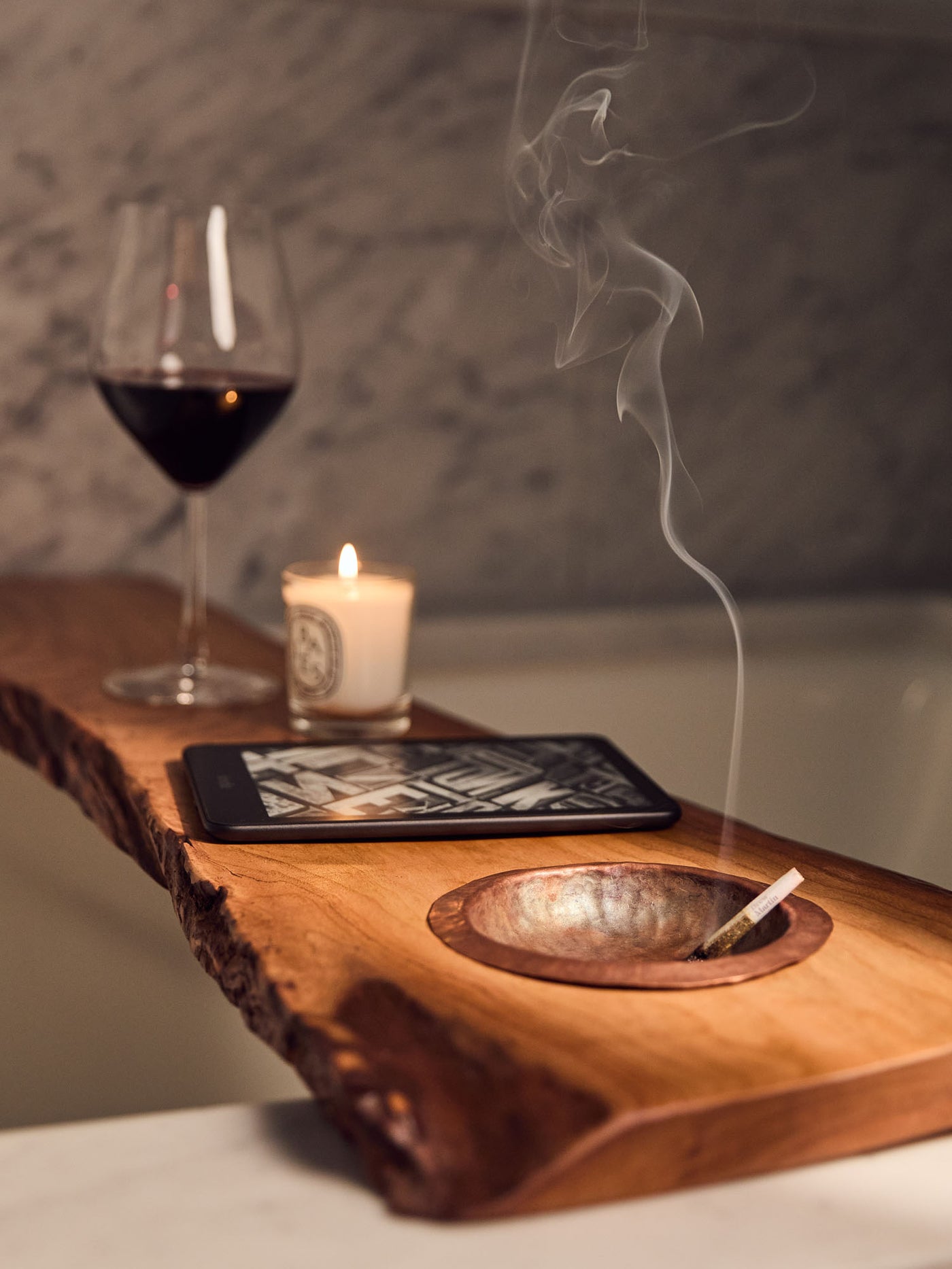  live-edge cherry wood bath tray with a hand-hammered copper ashtray over bathtub, with candle, wine glass and e-reader on it