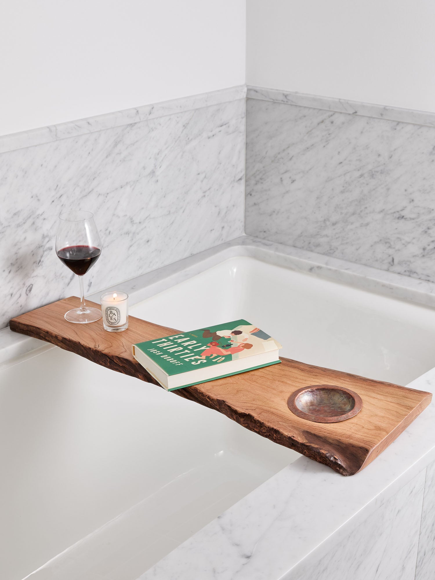 live-edge cherry wood bath tray with a hand-hammered copper ashtray over bathtub, with candle, wine glass and e-book on it