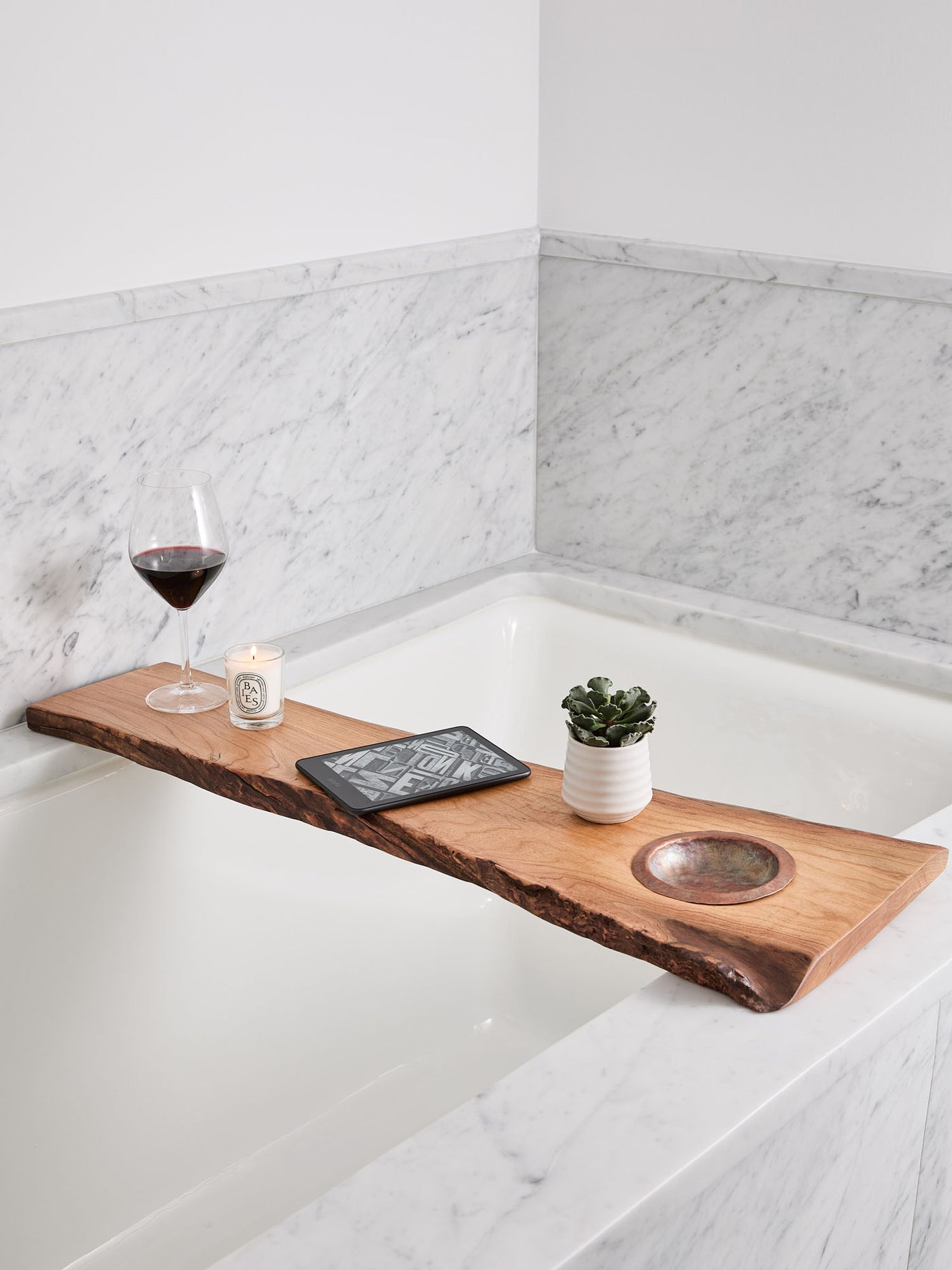  live-edge cherry wood bath tray with a hand-hammered copper ashtray over bathtub, with candle, wine glass, plant, and e-reader on it