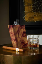 Cigar, glass of whiskey, and Judd wooden lighter on a reflective surface with abstract art in the background.