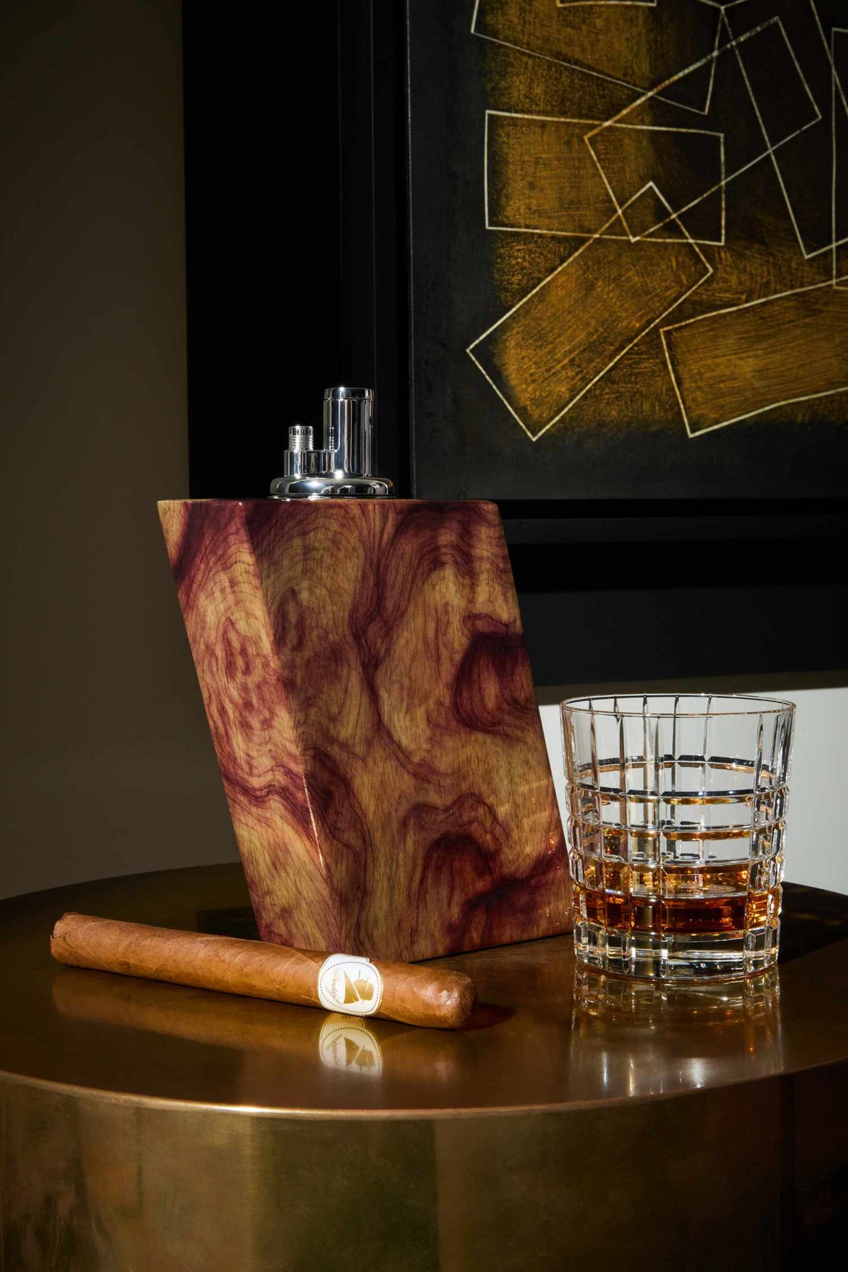 Cigar, glass of whiskey, and Judd wooden lighter on a reflective surface with abstract art in the background.