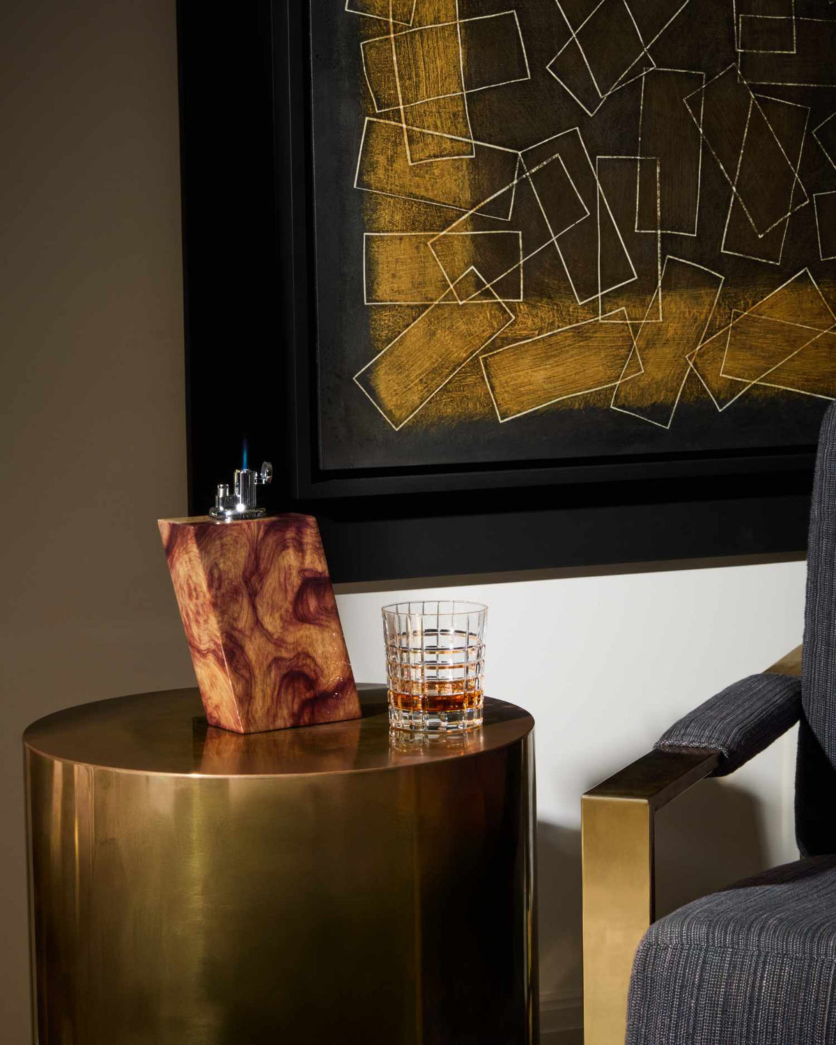 Gold side table with a glass of whiskey and wooden tabletop lighter, against a wall with abstract art.